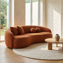 Enzo 89 Japandi curved sofa upholstered in burnt orange boucle fabric with matching throw pillows, styled in a minimalist sunlit living room on a round woven rug next to a low wooden coffee table.
