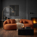 Modern minimalist living room featuring the Enzo 89 Japandi curved sofa in burnt orange boucle fabric, styled with matching square and spherical throw pillows and a beige waffle-knit throw blanket. In the foreground, a black marble block coffee table holds a lit candle. The cozy space is accented by a brass arched floor lamp, dark textured charcoal walls, and a glowing fireplace.