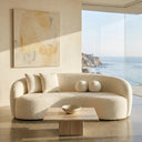 Enzo 89-inch Japandi ivory boucle curved sofa with matching throw pillows in a sunlit coastal living room, featuring a low travertine coffee table, neutral abstract wall art, and large floor-to-ceiling windows overlooking a scenic ocean and cliffside view.