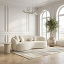 Enzo 89 Japandi ivory bouclé sofa in a bright, minimalist living room, featuring a curved design, spherical throw pillows, a fluted travertine side table, a patterned area rug on herringbone floors, and a potted olive tree by arched windows.