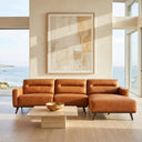 Erika 109-inch cognac PU leather right-facing sectional sofa with chaise and tapered wood legs in a sunlit modern living room overlooking the ocean, paired with a travertine coffee table and abstract wall art.