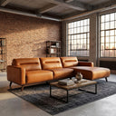 Erika 109-inch cognac brown PU leather right-facing sectional sofa with chaise lounge styled in an industrial loft living room with exposed brick walls, large factory grid windows, a patterned area rug, and a rustic wood coffee table.