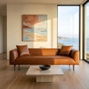 Erling 86-inch cognac vegan eco-leather sofa in a modern minimalist living room featuring a travertine stone coffee table, abstract coastal painting, and large floor-to-ceiling window overlooking a sunny ocean view.