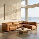 Fiorella 103" cognac brown vegan eco-leather modular corner sectional sofa in a sunlit modern living room with floor-to-ceiling ocean view windows and a low travertine coffee table.