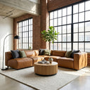 Fiorella 103-inch cognac vegan eco-leather corner sectional sofa styled with accent pillows and a round fluted wooden coffee table on a woven area rug in an industrial loft living room with exposed brick walls and large grid windows.