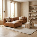 Fiorella 103-inch cognac vegan eco-leather corner sectional sofa with chaise and decorative pillows in a modern, sunlit living room with travertine nesting coffee tables and herringbone wood floors.