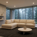 Gamila 110-inch beige genuine top-grain leather left-facing sectional sofa with chaise lounge in a modern, minimalist living room with floor-to-ceiling windows showing a misty forest view, set on a dark gray rug with a round stone coffee table.