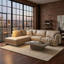 Gamila 110-inch beige genuine top-grain leather left-facing sectional sofa in a modern industrial loft living room with exposed brick walls, dark wood floors, a textured cream area rug, a concrete coffee table, and a large grid window showcasing a city skyline at dusk.