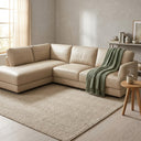 Gamila 110-inch beige genuine top-grain leather left-facing sectional sofa styled in a modern, neutral-toned living room. The L-shaped leather sectional features a soft sage green throw blanket draped over one end and is placed on a textured beige chevron area rug. A small round wooden side table sits next to it, with a window and a decorated console table in the background.