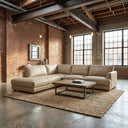Gamila 110 beige genuine top-grain leather left-facing sectional sofa in an industrial loft setting with exposed brick walls, large windows, a rustic wooden coffee table, and a jute area rug.