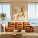 Gina 117 cognac genuine leather power slider sofa in a modern coastal living room with floor-to-ceiling windows showing ocean views, styled with a large abstract painting, travertine coffee table, and a potted olive tree.