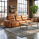 The Gina 117 cognac genuine leather power slider sofa in a modern loft living room with exposed brick walls, large industrial windows, a rustic wooden coffee table, a grey patterned rug, and a potted fiddle-leaf fig plant.