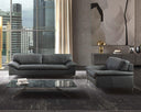 The Giorgia leather sofa and matching armchair in black, styled in a modern high-rise living room with a city skyline visible through large windows. The scene includes a glass coffee table, a textured grey rug, and large abstract art on the wall.