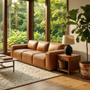 A Hanna 90-inch mid-century modern tan genuine leather sofa in a sunlit living room with floor-to-ceiling windows showing a lush garden. A walnut wood side table with a black ceramic table lamp and a potted fiddle leaf fig tree are on the right, all resting on a textured cream area rug over hardwood floors.