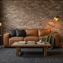 Hanna 90-inch mid-century modern tan genuine leather sofa against an exposed brick wall, styled with charcoal pillows, an olive green knit blanket, a rustic wood coffee table, a dark textured rug, and a warm brass floor lamp.