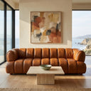 Mid-century modern Jess 90-inch cognac genuine leather sofa with channel tufting in a bright coastal living room, featuring a square travertine coffee table and abstract wall art with scenic ocean and cliff views through floor-to-ceiling windows.
