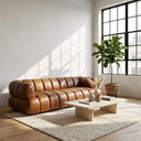 Jess 90-inch Mid-Century Modern cognac genuine leather channel-tufted sofa in a minimalist living room featuring a travertine coffee table, potted fiddle-leaf fig tree, and natural sunlight.