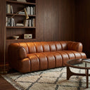 Jess 90-inch mid-century modern cognac genuine leather sofa with channel tufting, styled in a living room with dark wood wall paneling, a built-in bookshelf, a brass arched floor lamp, a patterned area rug, and a glass-top coffee table.
