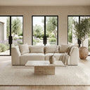 The June 98 modern beige chenille sofa with matching cushions and a light throw blanket in a minimalist living room. A travertine coffee table sits on a textured cream area rug, with large black-framed windows showing a garden background.