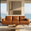 Kaia 98-inch tan genuine leather sofa in a modern coastal living room with floor-to-ceiling windows overlooking the ocean, featuring a stone coffee table and large abstract artwork.