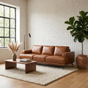 Kaia 98-inch tan genuine leather sofa in a modern living room with a white brick wall, a low wooden coffee table, a textured cream rug, and a large fiddle-leaf fig plant.