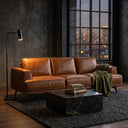 Kaia 98-inch tan genuine leather sofa in a modern industrial living room, styled with an olive green throw blanket, featuring a black marble coffee table, dark textured walls, and a large window showing city lights at night.