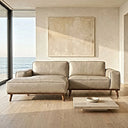 Beige leather left-facing sectional sofa with contrast piping and tapered wooden legs in a sunlit coastal living room with light wood floors, a low stone coffee table, neutral canvas art, and floor-to-ceiling ocean-view windows.