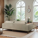 Back angle view of the Katia 98-inch beige leather left-facing sectional sofa with a wooden base and angled legs, placed on a textured rug in a sunlit living room with arched windows, indoor palms, and a hanging wall clock.
