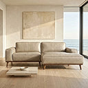 Beige leather right-facing sectional sofa with contrast piping and tapered wooden legs in a sunlit coastal living room with light wood floors, a low stone coffee table, neutral canvas art, and floor-to-ceiling ocean-view windows.
