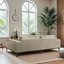 Back angle view of the Katia 98-inch beige leather right-facing sectional sofa with a wooden base and angled legs, placed on a textured rug in a sunlit living room with arched windows, indoor palms, and a hanging wall clock.
