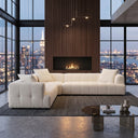 Kendall 120-inch ivory boucle L-shaped corner sofa with channel-tufted cushions and matching throw pillows, set on a dark woven rug in a modern high-rise loft with a dark wood slat fireplace wall and floor-to-ceiling city skyline windows.