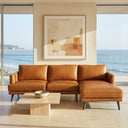 Lara 106-inch L-shaped right-facing sectional sofa in genuine tan leather with tapered dark wood legs, styled in a sunlit modern living room with a travertine coffee table and floor-to-ceiling ocean view windows.