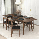 Mid-century modern rectangular walnut dining table with angled legs, surrounded by six matching wood chairs with black cushions, set with six black plates and a dried floral centerpiece on a textured beige rug in a bright dining room.
