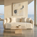 Lina 84 Ivory Boucle Sofa in a modern, sunlit living room with a travertine stone coffee table, abstract wall art, and scenic coastal ocean views through large windows.