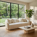 Lina 84-inch ivory boucle sofa with a curved silhouette and matching throw pillows in a bright, modern living room with light oak herringbone floors, a travertine block coffee table, a potted fiddle-leaf fig tree, and large black-framed windows overlooking a lush green garden.