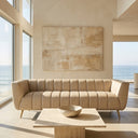 Lorena 86-inch beige genuine Italian leather sofa with vertical channel tufting and gold legs in a modern minimalist living room overlooking the ocean, featuring a travertine coffee table and abstract canvas art.