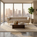 Lorena 86" beige genuine Italian leather sofa with vertical channel tufting and gold legs, styled in a modern living room with a wood coffee table and floor-to-ceiling windows overlooking a city skyline at sunset.