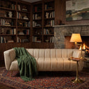 Lorena 86-inch beige genuine Italian leather sofa with vertical channel tufting, styled with a green throw blanket in a cozy living room featuring a stone fireplace and built-in bookshelves.