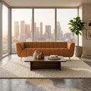 Lorena 86" beige genuine Italian leather sofa with vertical channel tufting and gold legs, styled in a modern living room with a wood coffee table and floor-to-ceiling windows overlooking a city skyline at sunset.