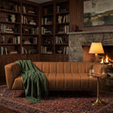 Lorena 86-inch cognac genuine Italian leather sofa with vertical channel tufting, styled with a green throw blanket in a cozy living room featuring a stone fireplace and built-in bookshelves.