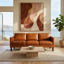 Mid-century modern cognac leather sofa in a bright, minimalist living room with an ocean view, styled with a travertine coffee table, large abstract wall art in warm earth tones, and a potted fiddle-leaf fig plant.
