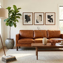 Mid-century modern living room featuring a three-seater cognac brown leather sofa, a wooden coffee table, a potted fiddle leaf fig tree, and three framed abstract art prints hanging on the wall.