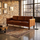Mid-century modern cognac leather sofa in a loft-style living room with an exposed brick wall, patterned rug, and large window.