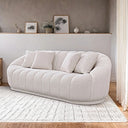 Curved white French bouclé sofa with vertical channel tufting and four matching square throw pillows, resting on a textured cream rug in front of a neutral two-tone wall with a decorative ledge.
