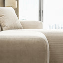 Close-up detail of the Melina cream corduroy sectional sofa, showcasing the textured ribbed upholstery on the plush cushion, armrest, and matching pillow beside a bright window with a city skyline view.