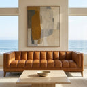Mia 90 mid-century modern cognac vegan eco-leather sofa with vertical channel tufting in a bright living room featuring a travertine coffee table, abstract painting, and ocean view.