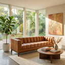 Mia 90-inch mid-century modern cognac vegan eco-leather sofa with vertical channel tufting in a bright living room, featuring a round wooden coffee table, a potted fiddle-leaf fig, and large floor-to-ceiling windows.
