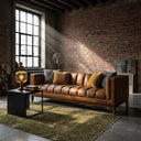 Mia 90-inch mid-century modern cognac vegan eco-leather sofa with channeled tufting, styled in an industrial loft living room with an exposed brick wall, green area rug, and amber table lamp.