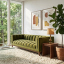 Mia 90 Mid-Century Modern olive green velvet sofa with channel tufting and a wooden base in a sunlit living room, featuring a cream shag rug, a walnut side table with a gold lamp, abstract framed art, and a potted fiddle-leaf fig.