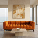 Mia 90 mid-century modern orange velvet sofa with vertical channel tufting in a bright living room, featuring a low travertine coffee table, large abstract wall art, and floor-to-ceiling windows with a scenic ocean view.
